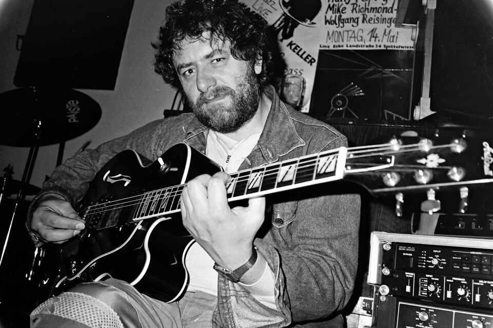 Schwarzweißporträt: Harry Pepl sitzt mit einer Jazzgitarre im Arm und blickt in die Kamera; an der Wand das Plakat eines Konzerts mit Mike Richmond und Wolfgang Reisinger in Linz („Montag, 14. Mai“), rechts ein Effektgerät.